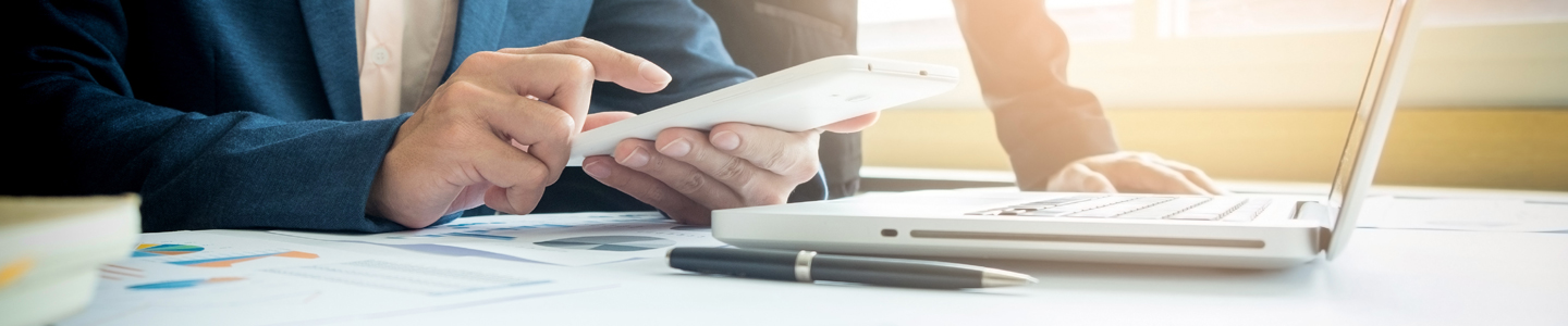 M�os de um homem mexendo no celular, a sua frente est�o um laptop, uma caneta e pap�is com gr�ficos impressos.